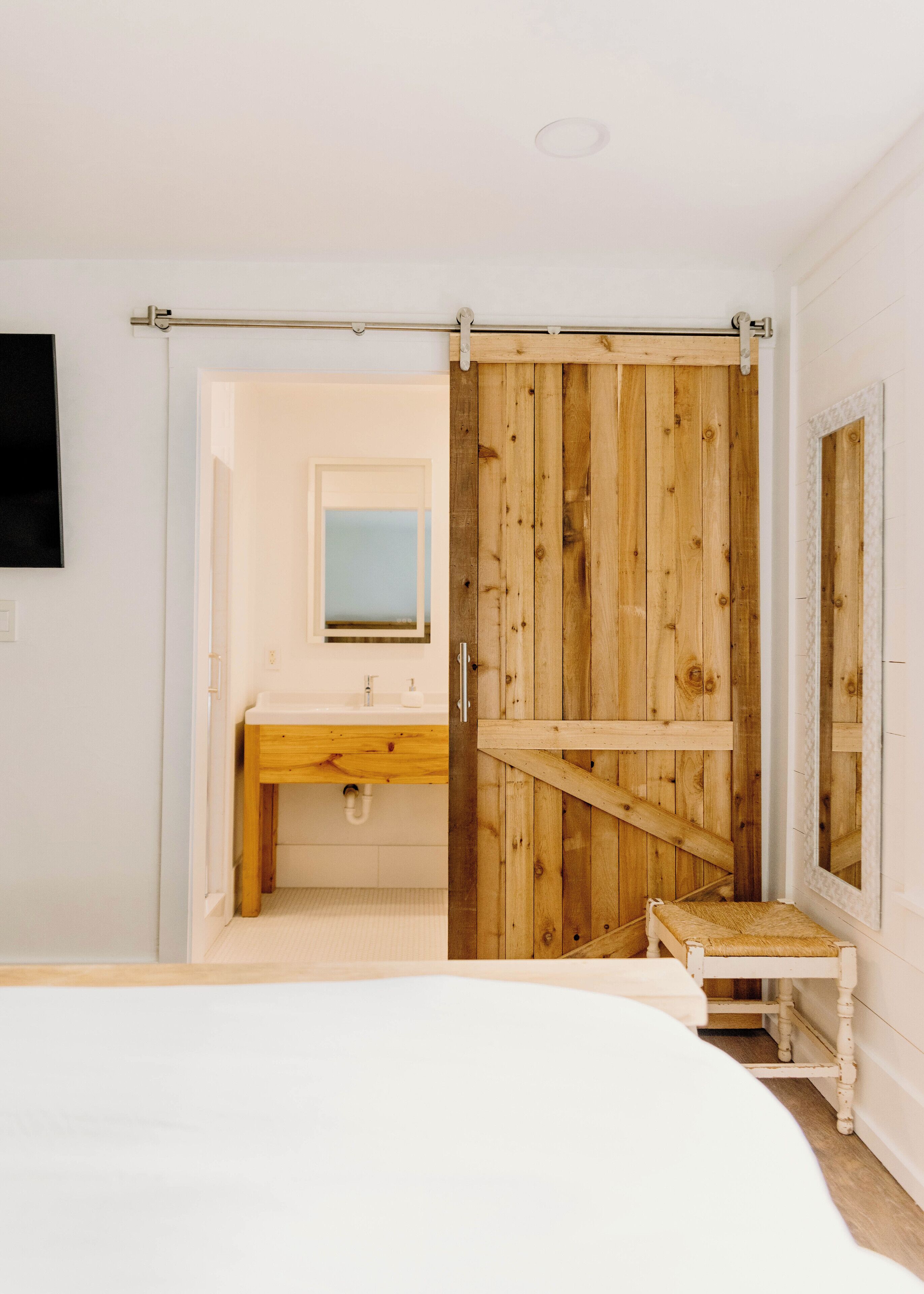 Un dormitorio con cama, un armario de madera, un baño con lavamanos y espejo, y un taburete pequeño.