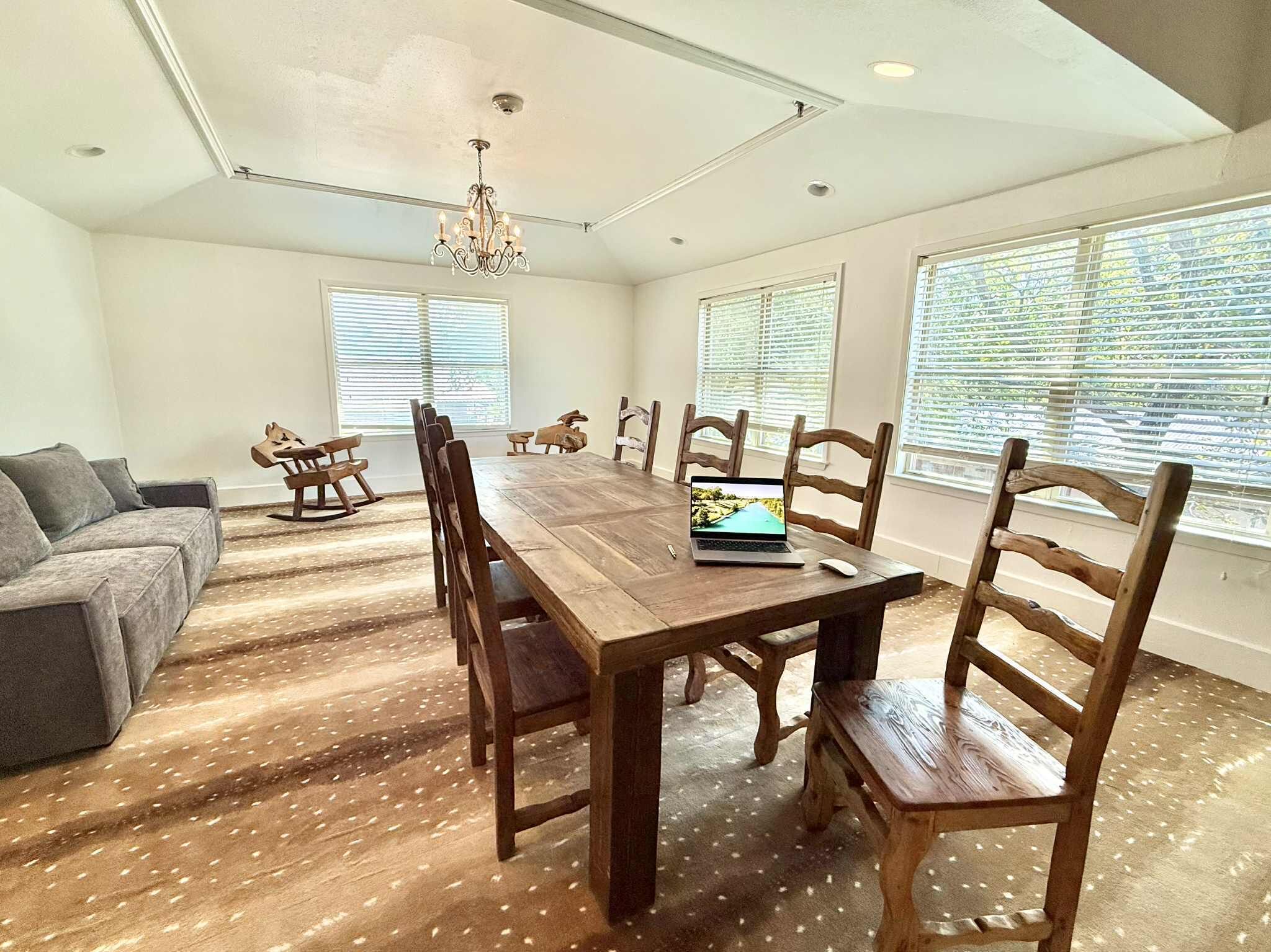 A spacious room with a large wooden dining table, six chairs, a grey sofa, and a chandelier.