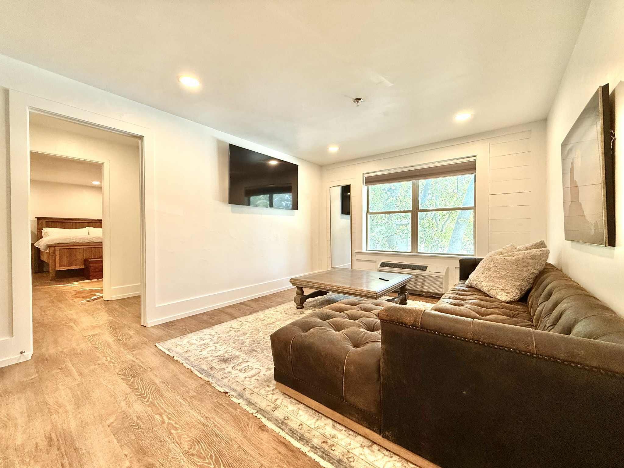 A spacious living room with a brown leather sofa, a wooden coffee table, and a large window providing natural light.