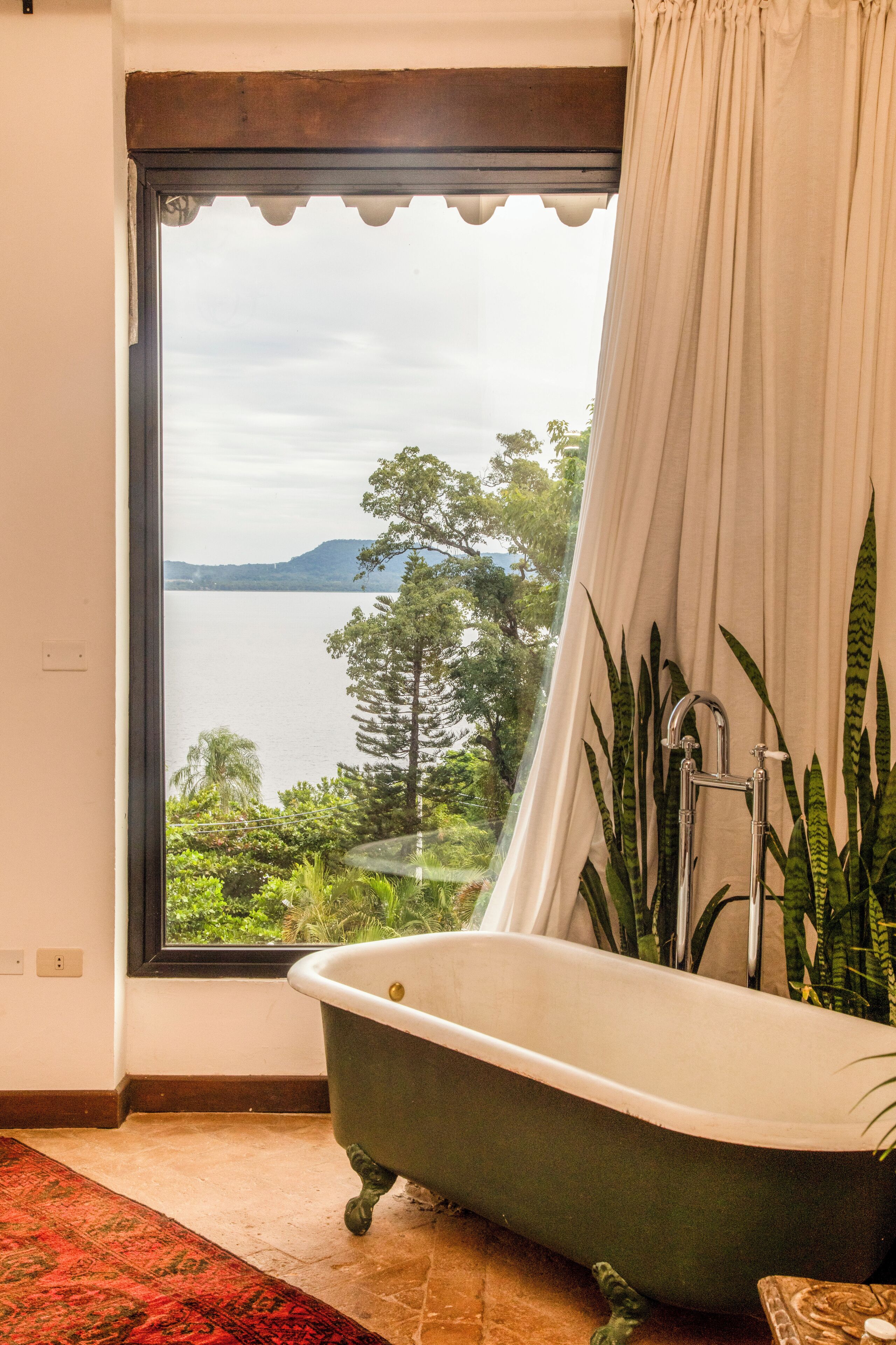 Un baño con bañera, un ventanal con vistas a un lago y árboles, y una cortina.