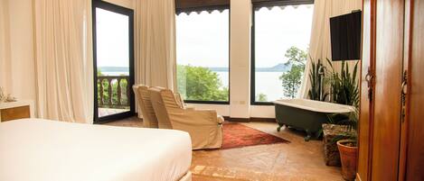 A bedroom with a large bed, a chaise lounge, a bathtub, and a view of the outdoors.