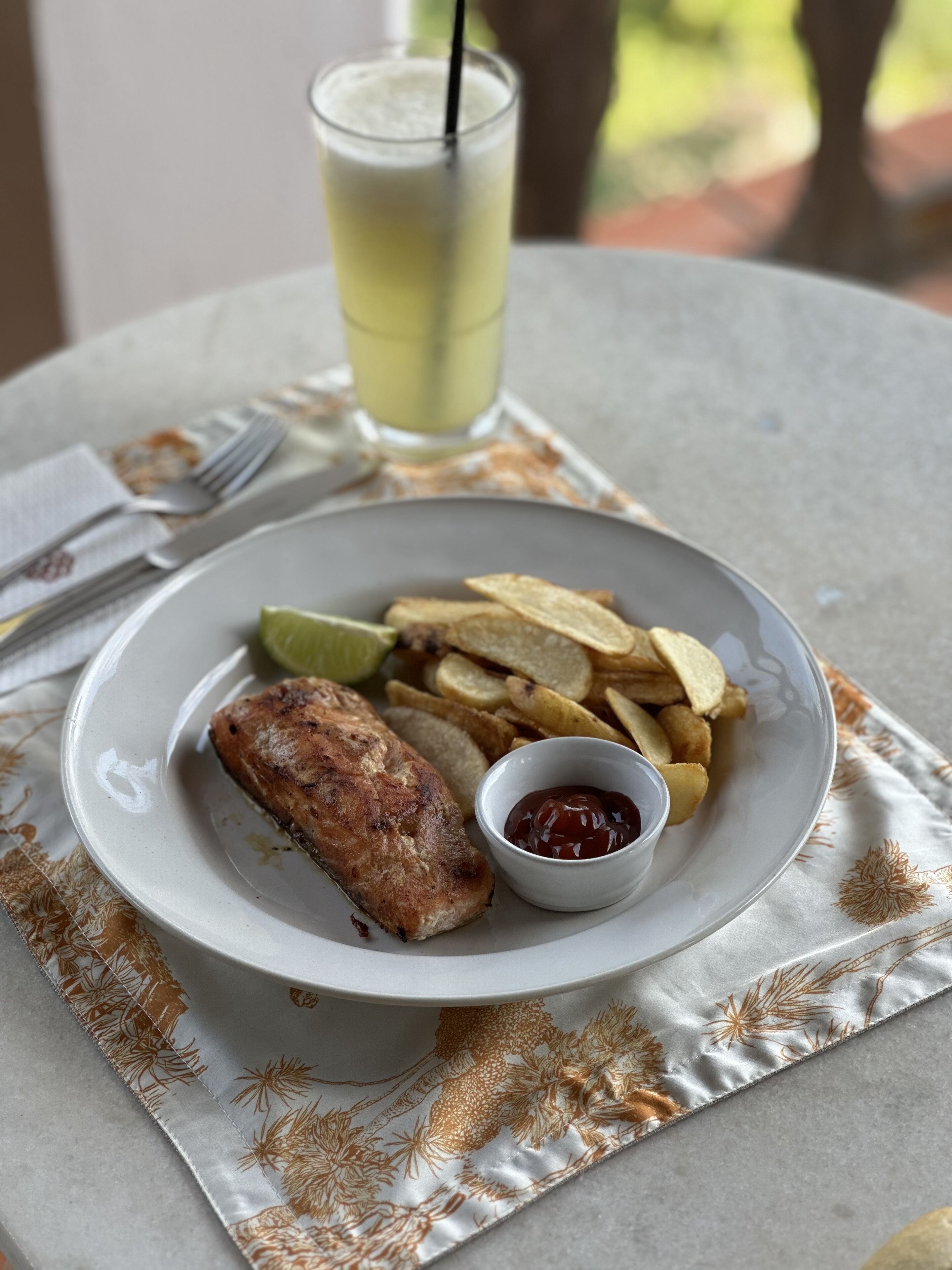 A plate with a grilled fish, potato chips, and a small bowl of ketchup, accompanied by a glass of a yellow beverage on a table with a patterned tablecloth.