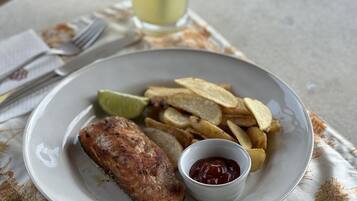 A plate with a grilled fish, potato chips, and a small bowl of ketchup, accompanied by a glass of a yellow beverage on a table with a patterned tablecloth.
