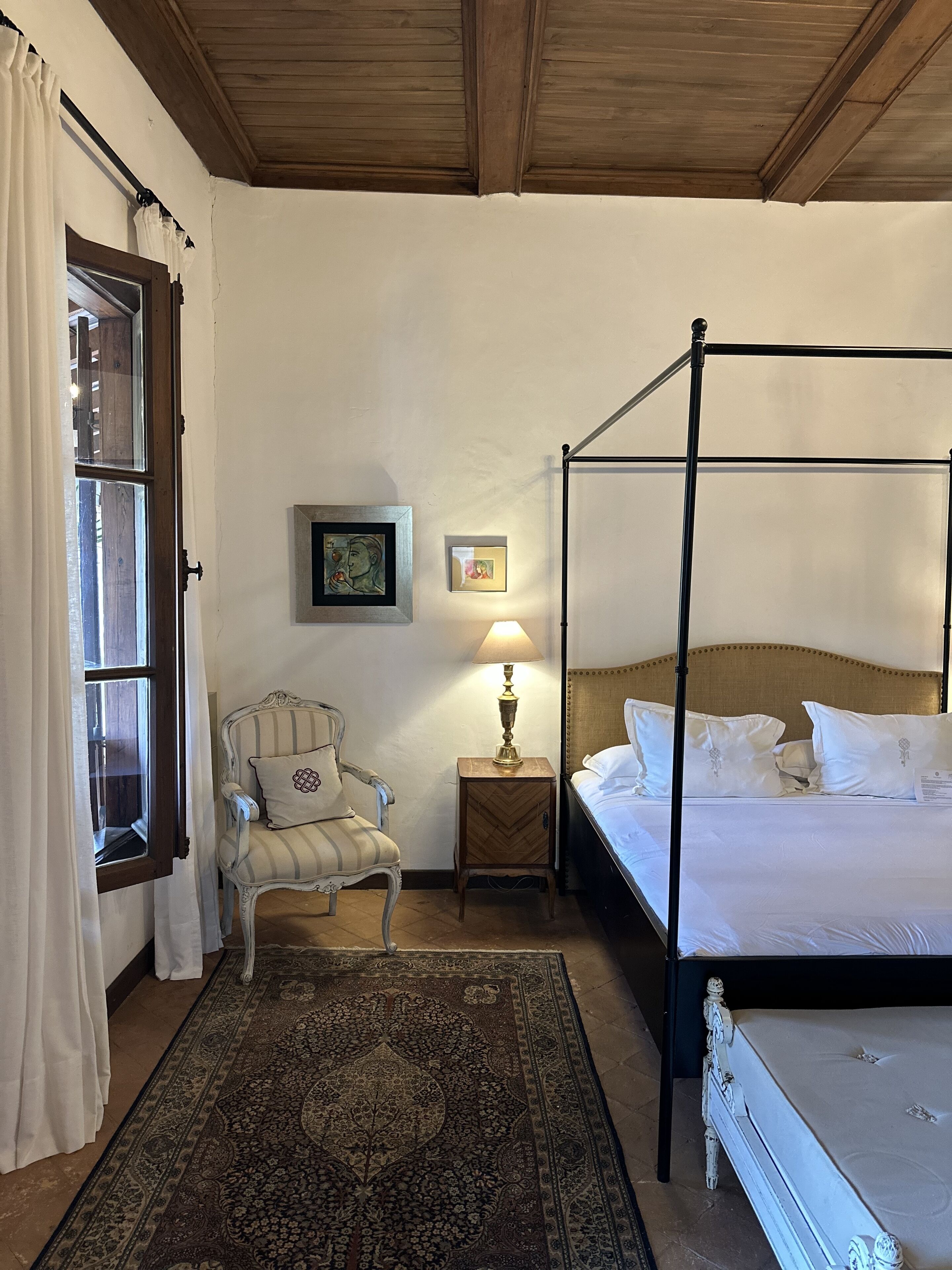 A four-poster bed with white linens, a small bedside table with a lamp, a chair with striped upholstery, and a patterned rug.