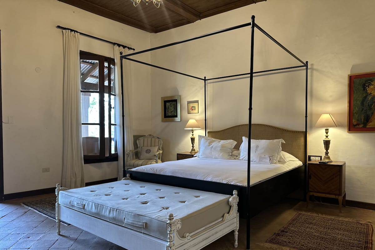 Una cama con dosel, colchón blanco y cabecera, dos lámparas de noche, una silla y una alfombra en una habitación con suelo de baldosas y una ventana con cortinas.