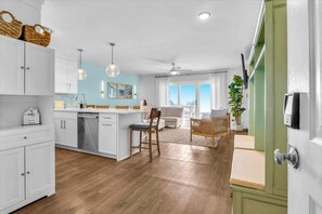 A modern kitchen with white cabinetry, a central island, pendant lighting, and a view of the living area with a sofa and a plant.