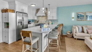 A modern kitchen with white cabinets, a stainless steel refrigerator, a central island with bar stools, and a light blue wall. - Oceanfront 2BR Suite | Boardwalk-Parking-Sleeps 5 (Carolina Beach)