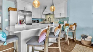 A modern kitchen with white cabinetry, a central island, two pendant lights, and bar stools with cushioned seats. - Oceanfront 2BR Suite | Boardwalk-Parking-Sleeps 5 (Carolina Beach)