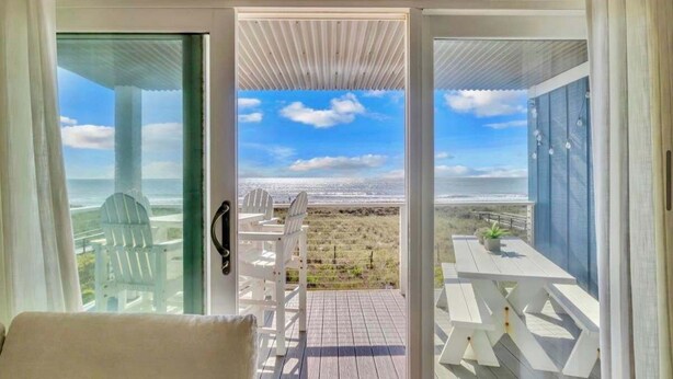 A room with a view of the beach, a white chair, a table, and a door leading outside. - Oceanfront 2BR Suite | Boardwalk-Parking-Sleeps 5 (Carolina Beach)