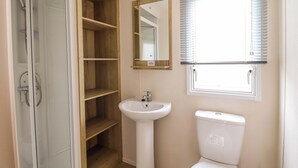 A bathroom with a glass shower enclosure, a pedestal sink, a toilet, wooden shelving, a mirror, and a window with blinds. - Beachcombers Barmouth Bay Holiday Park (Talybont)