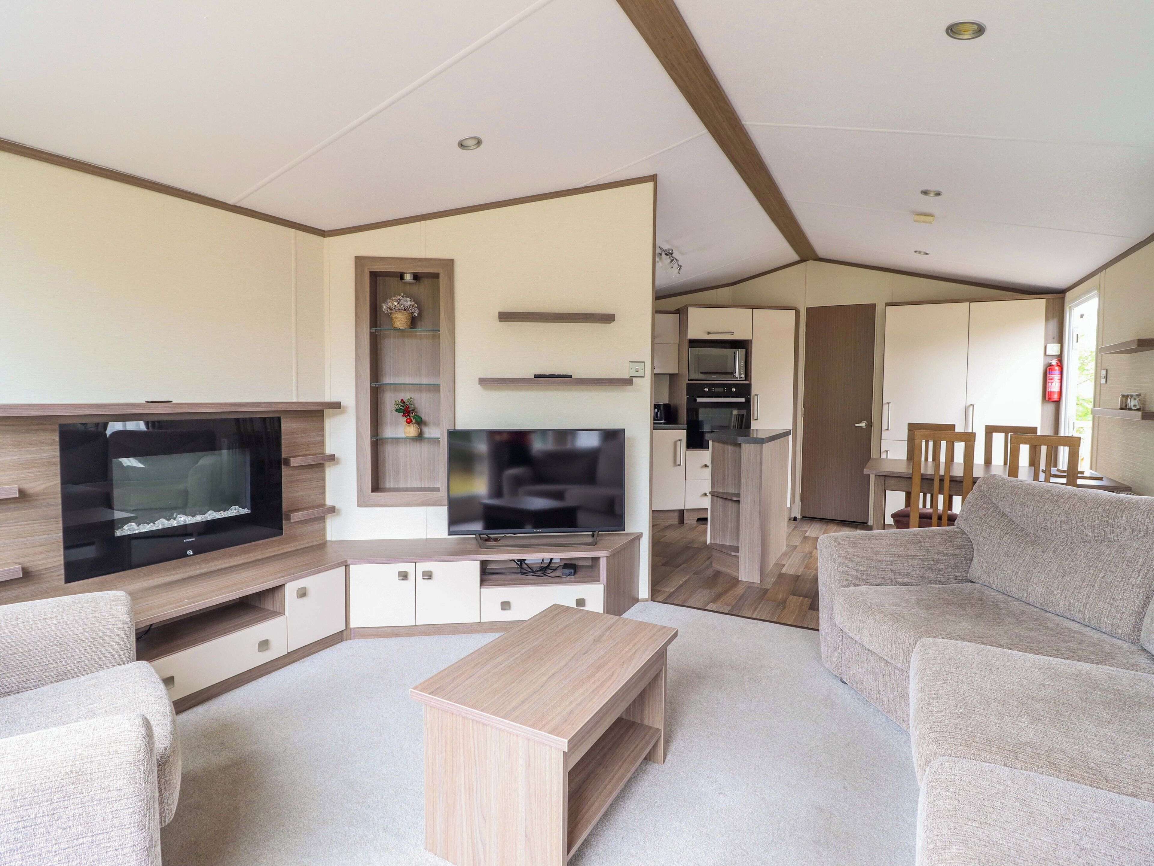 A modern living room with a flat-screen TV, a wooden coffee table, and a sofa. There is a kitchen area with a microwave and oven in the background.