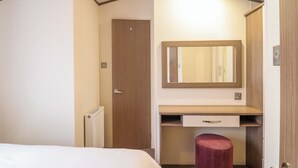 A compact hotel room with a bed, a small desk with a mirror, a red stool, and a wardrobe.