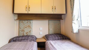 A small, compact bedroom with two single beds, a wooden wardrobe, and a window with curtains. - Sea Breeze C38 Dyffryn Seaside Estate (Dyffryn Ardudwy)