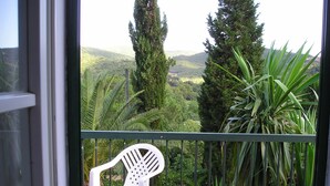 Un balcón con una silla de plástico blanca, barandilla metálica y vistas de un paisaje verde exuberante con árboles y colinas.