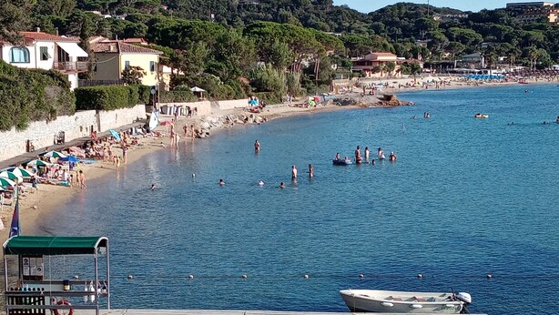 Zona costera con muelle, barcos y playa con sombrillas y gente.