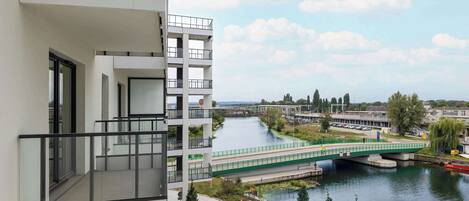 Un edificio moderno con balconi affacciati su un fiume, un ponte pedonale e un'area verde con alberi.