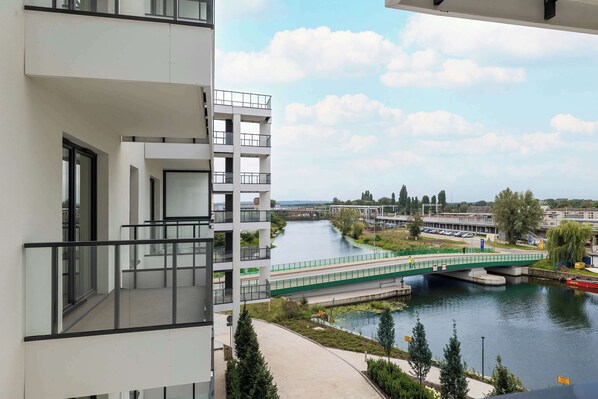 A modern building with balconies overlooking a river, a pedestrian bridge, and a park area with trees.
