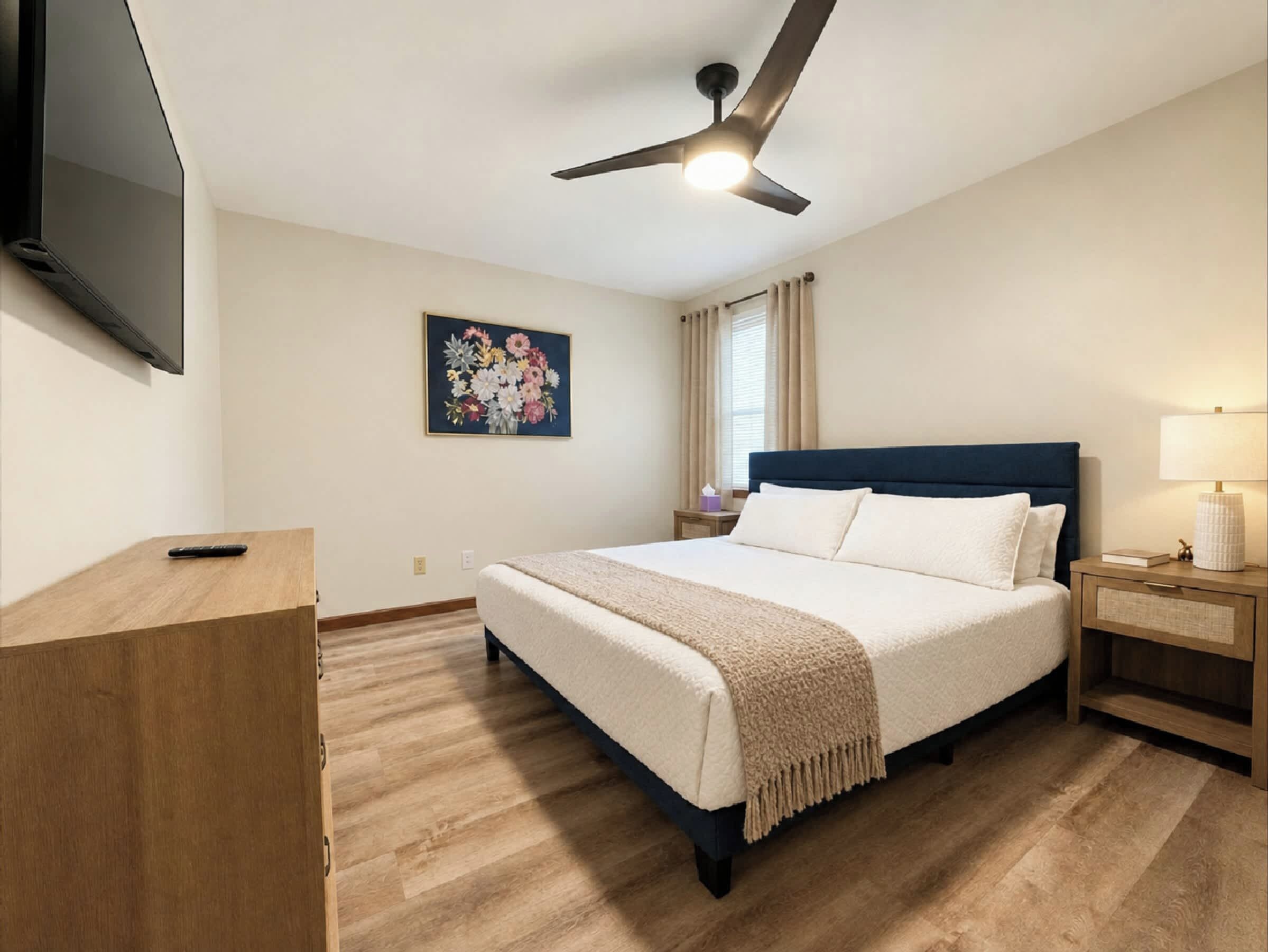 Relax in this spacious primary bedroom featuring a comfortable bed, smart TV, ceiling fan, and stylish furnishings. A peaceful retreat for a restful night's sleep after exploring Downtown Hamilton, Ohio.