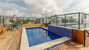 A rooftop pool area with a blue-tiled pool, wooden deck, and outdoor seating. - Aptos completos em em prédio a 400m da praia \- PZO (Ilhéus)