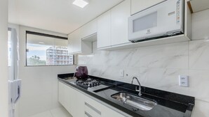 A modern kitchen with white cabinets, a black countertop, a built-in microwave, and a window with a view of a building. - Aptos completos em em prédio a 400m da praia \- PZO (Ilhéus)