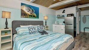 A bedroom with a bed, bedside tables, a ceiling fan, a painting, a kitchenette, and a refrigerator. - Waterfront Coconut Cottage in Fort Myers Beach (Fort Myers Beach)