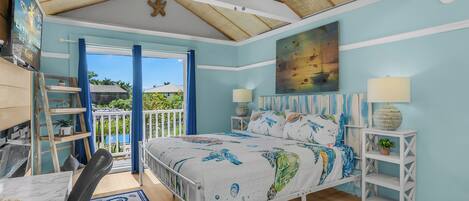 A bedroom with a bed, two bedside tables, a ceiling fan, a large window, and a view of the outdoors.