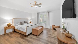 A modern bedroom with a wooden bed, bedside tables, a bench, a ceiling fan, and a TV. - The DOVI Collection | Cozy Forest Hideaway (The Woodlands)