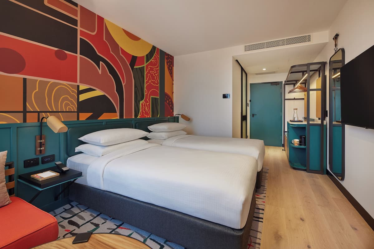 A hotel room with a large bed, a desk with a lamp, a red sofa, and a colorful abstract wall.
