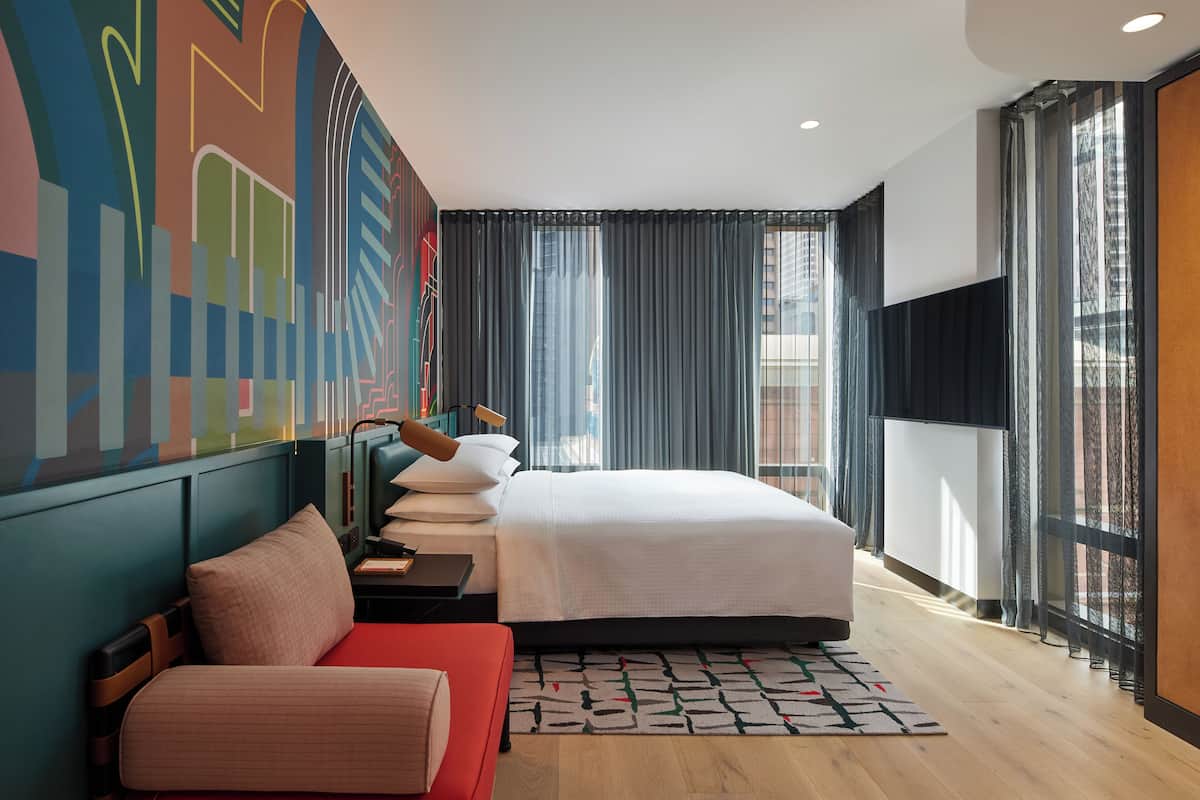A modern hotel room with a large bed, a red sofa, a desk with a lamp, and a colorful abstract mural on the wall.