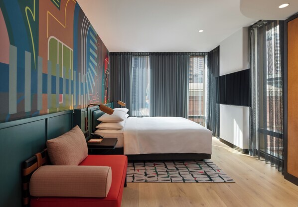 A modern hotel room with a large bed, a red sofa, a desk with a lamp, and a colorful abstract mural on the wall. - Caption By Hyatt Central Sydney (Haymarket)