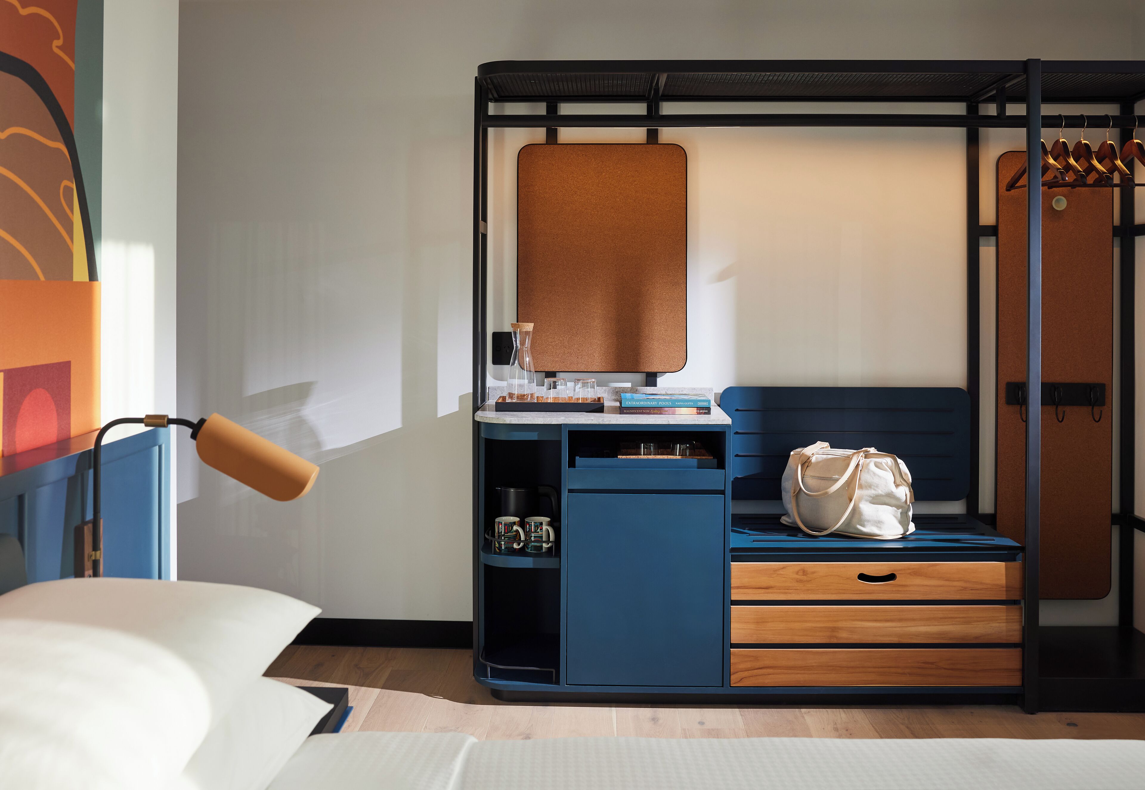 Une pièce de taille modeste comprenant un lit, un bureau avec un ordinateur, une chaise, un porte-manteau mural et un petit meuble de rangement.