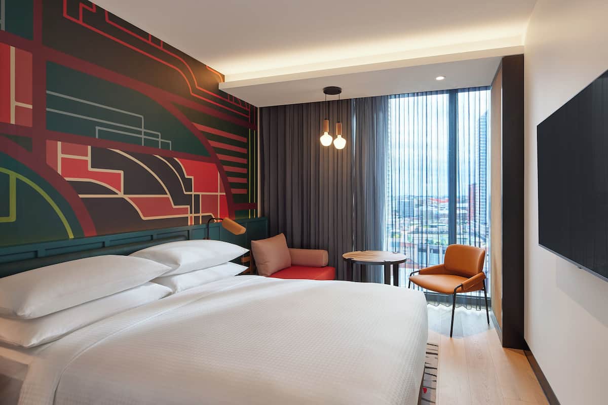 A modern hotel room with a large bed, a small seating area, and a view of the city through large windows.