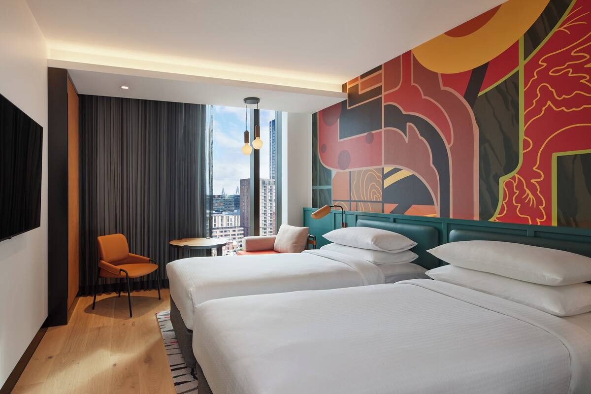 A hotel room with two beds, a chair, a small table, and a large window with a city view.