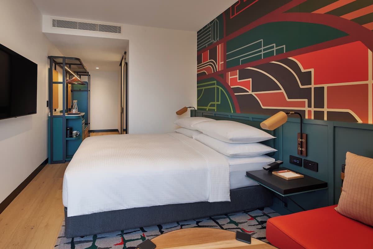A hotel room with a large bed, a desk with a lamp, a TV, and a colorful abstract wall mural.