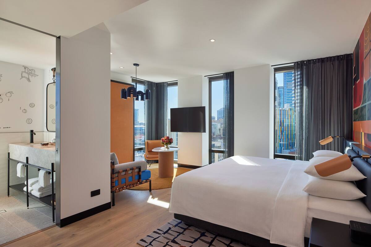 A modern hotel room with a large bed, a sitting area with a chair and table, a bathroom with a mirror and sink, and a city view through large windows.