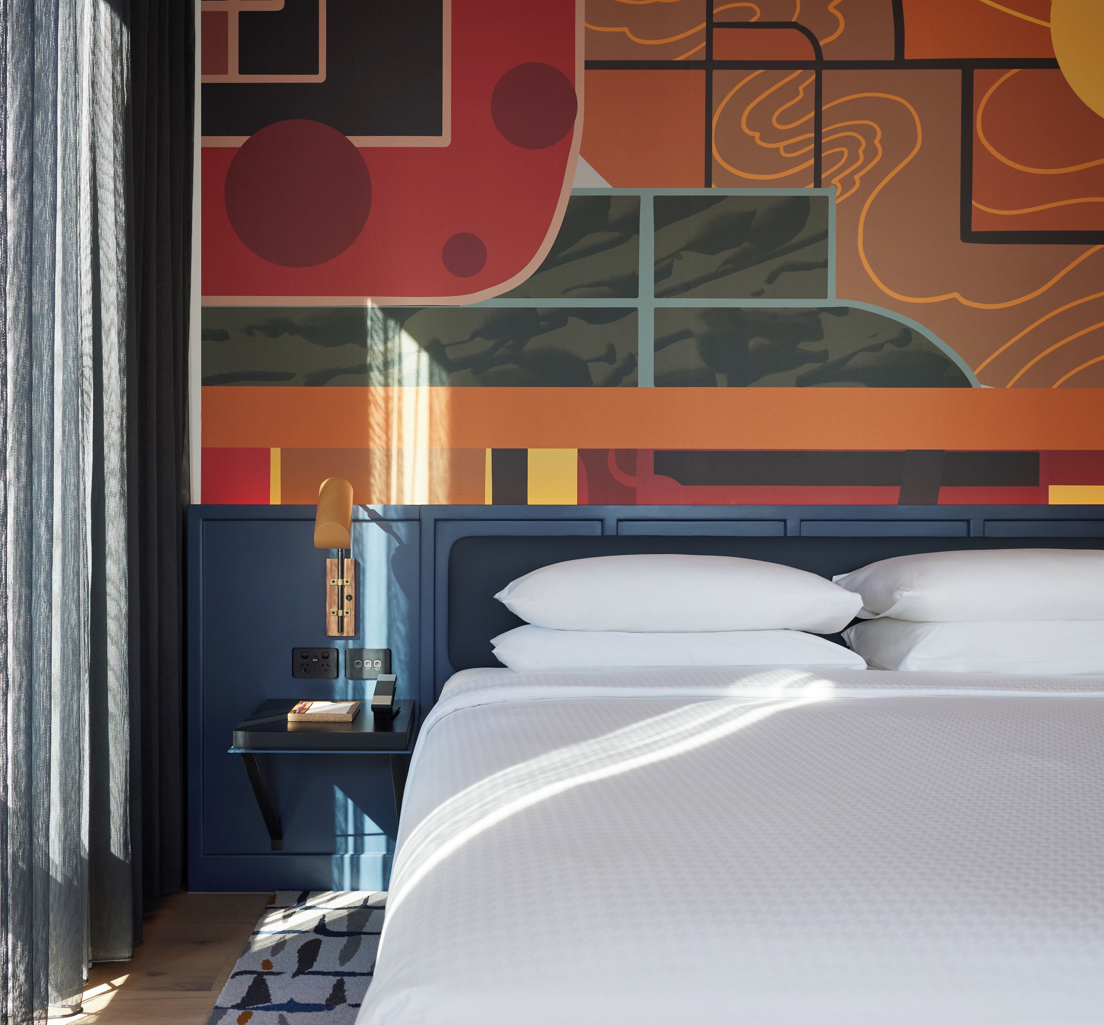 A modern hotel room with a large bed, a nightstand with a lamp, and a wall with abstract art.