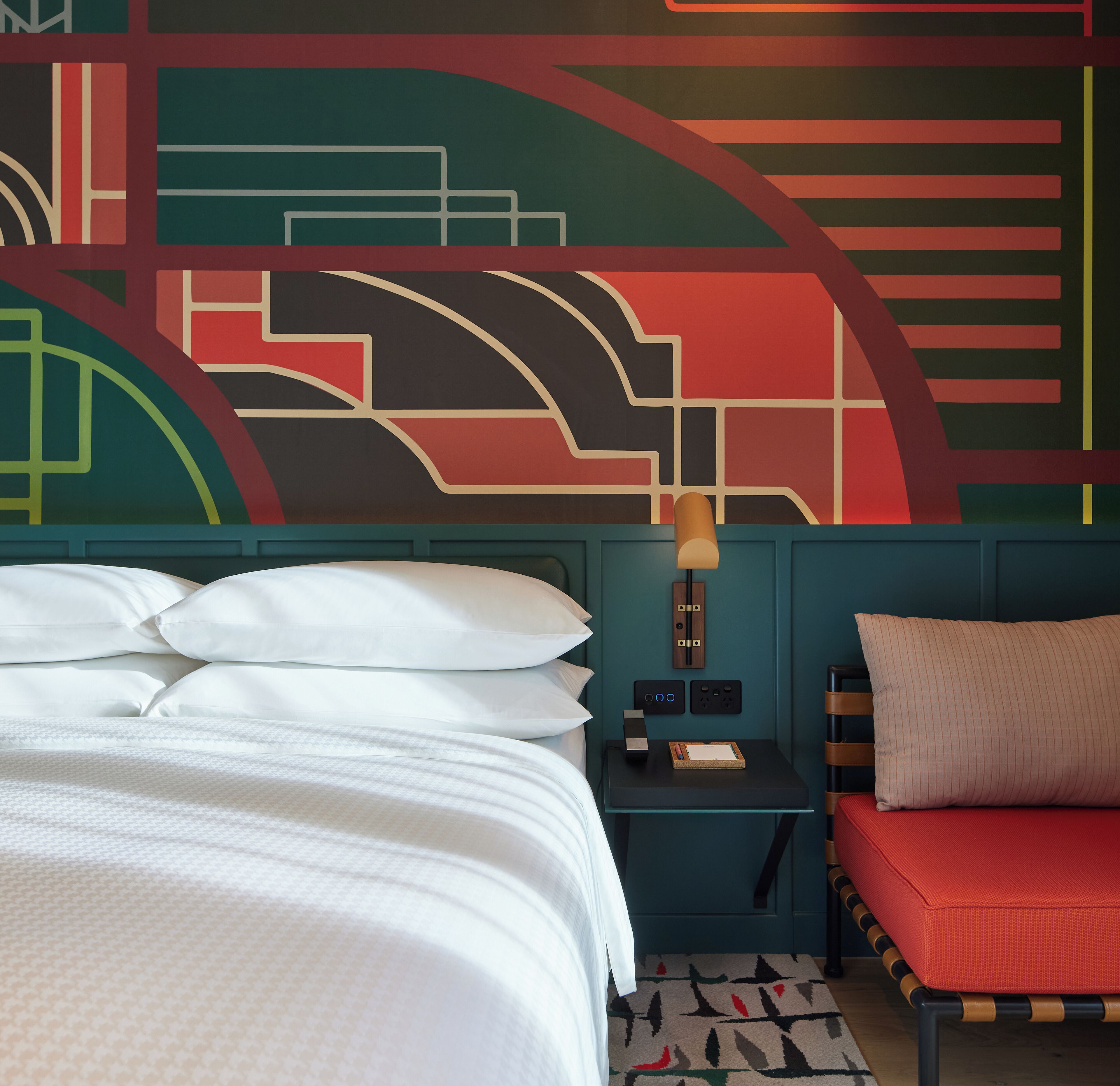 Habitación de hotel con cama, mesita de noche, lámpara, una silla roja y pared con diseños geométricos.