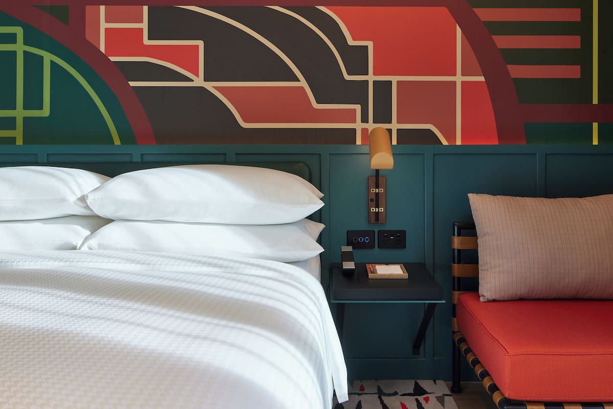 A hotel room with a bed, a nightstand, a lamp, a red chair, and a wall with geometric patterns.