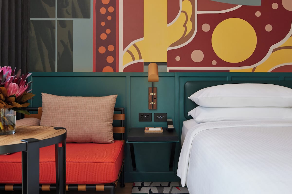 A hotel room with a bed, a nightstand, a table with a vase of flowers, and a colorful abstract wall mural.