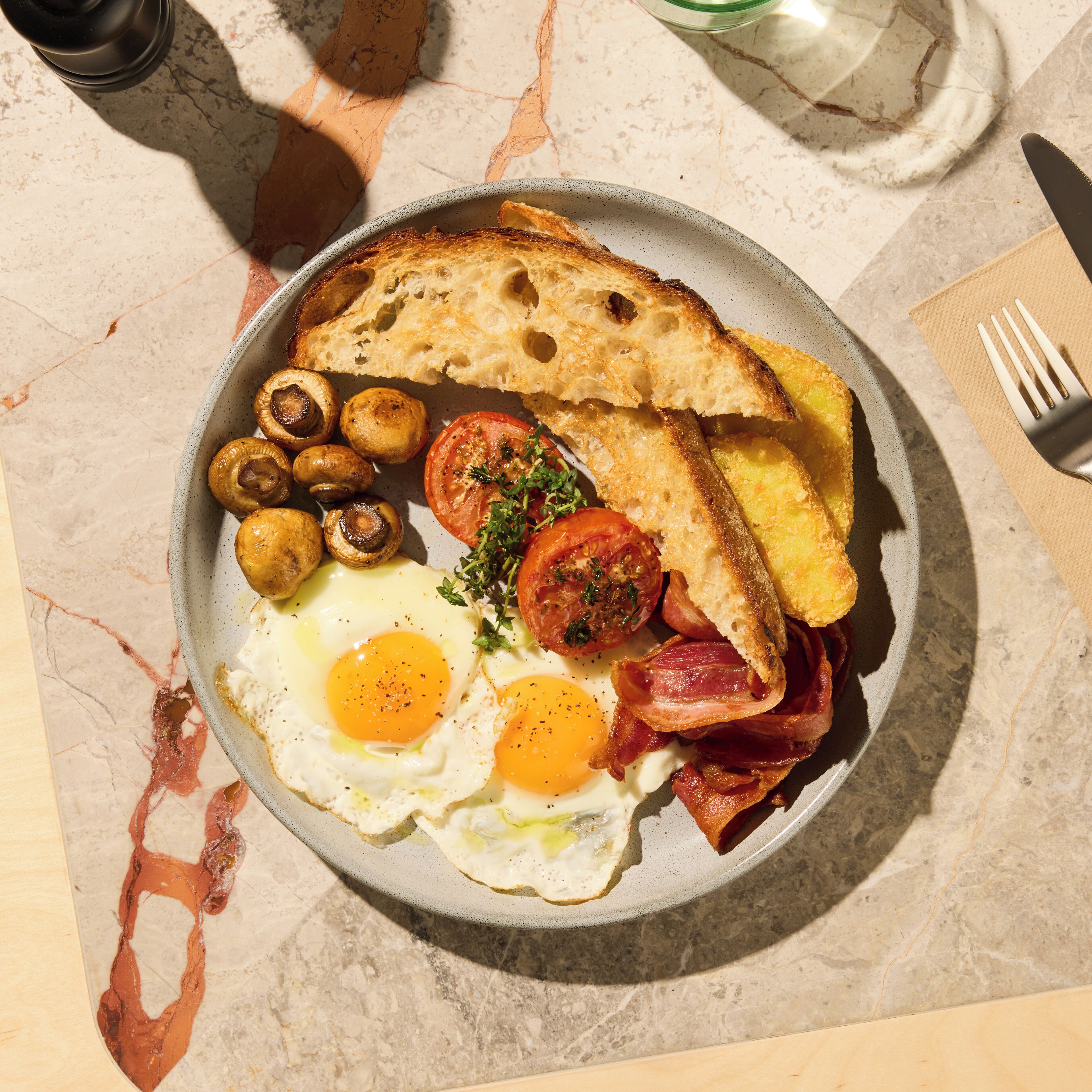A breakfast plate with eggs, bacon, mushrooms, tomatoes, and toast on a marble table.