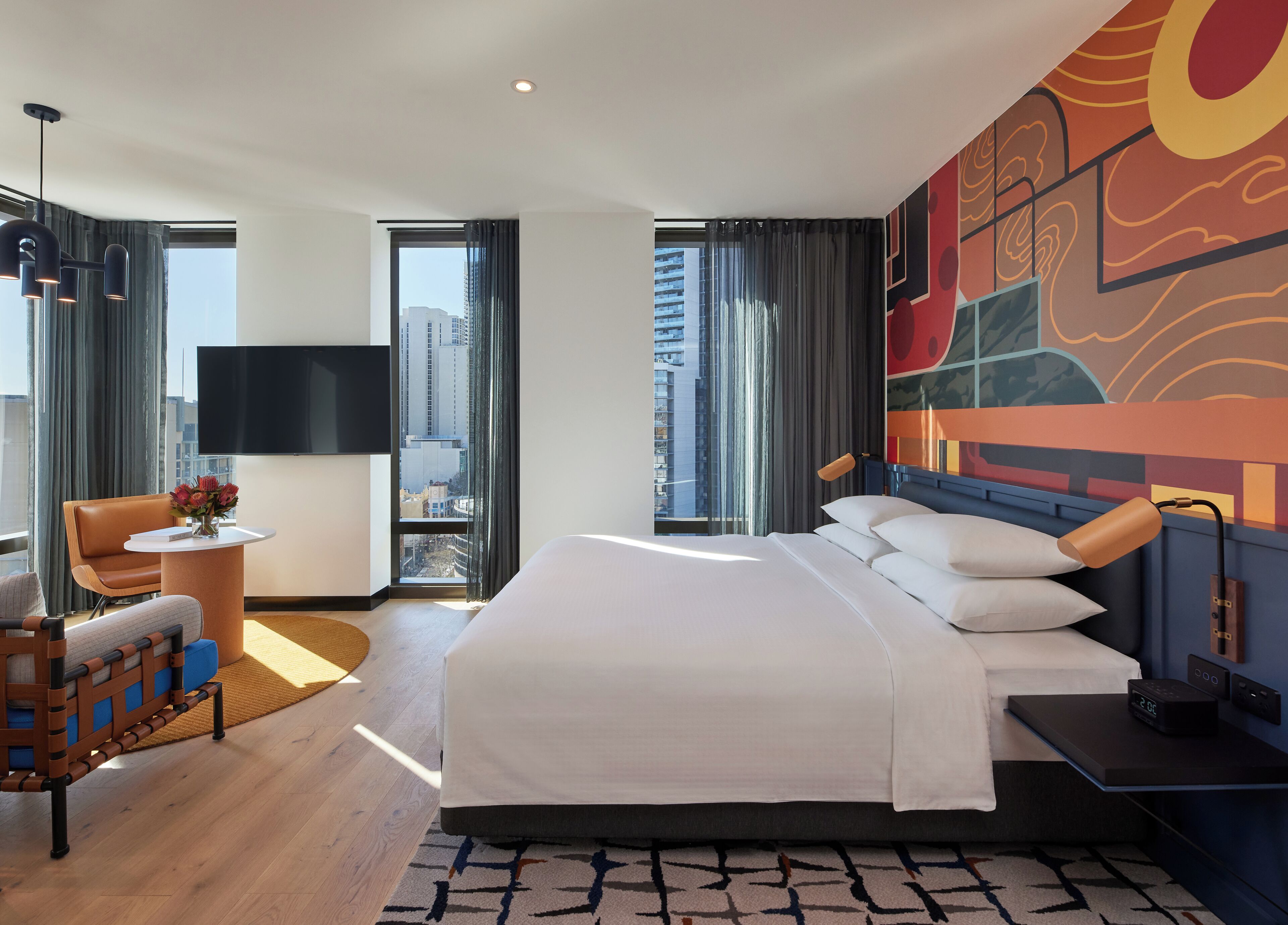 A modern hotel room with a large bed, a desk, a chair, a TV, and a city view.
