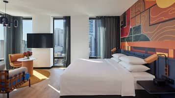 A modern hotel room with a large bed, a desk, a chair, a TV, and a city view.