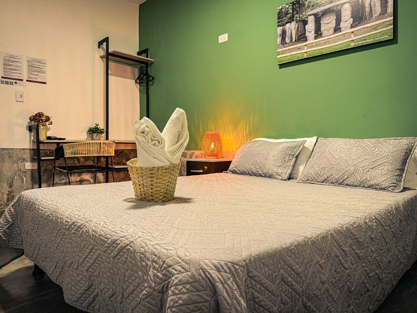 A neatly made bed with a quilted blanket, a wicker basket with folded towels, a small table with a lamp, and a framed picture on the wall.