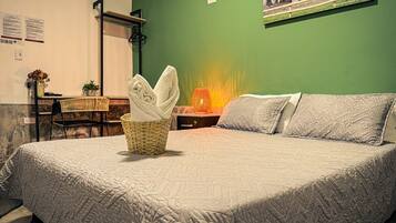 A neatly made bed with a quilted blanket, a wicker basket with folded towels, a small table with a lamp, and a framed picture on the wall.