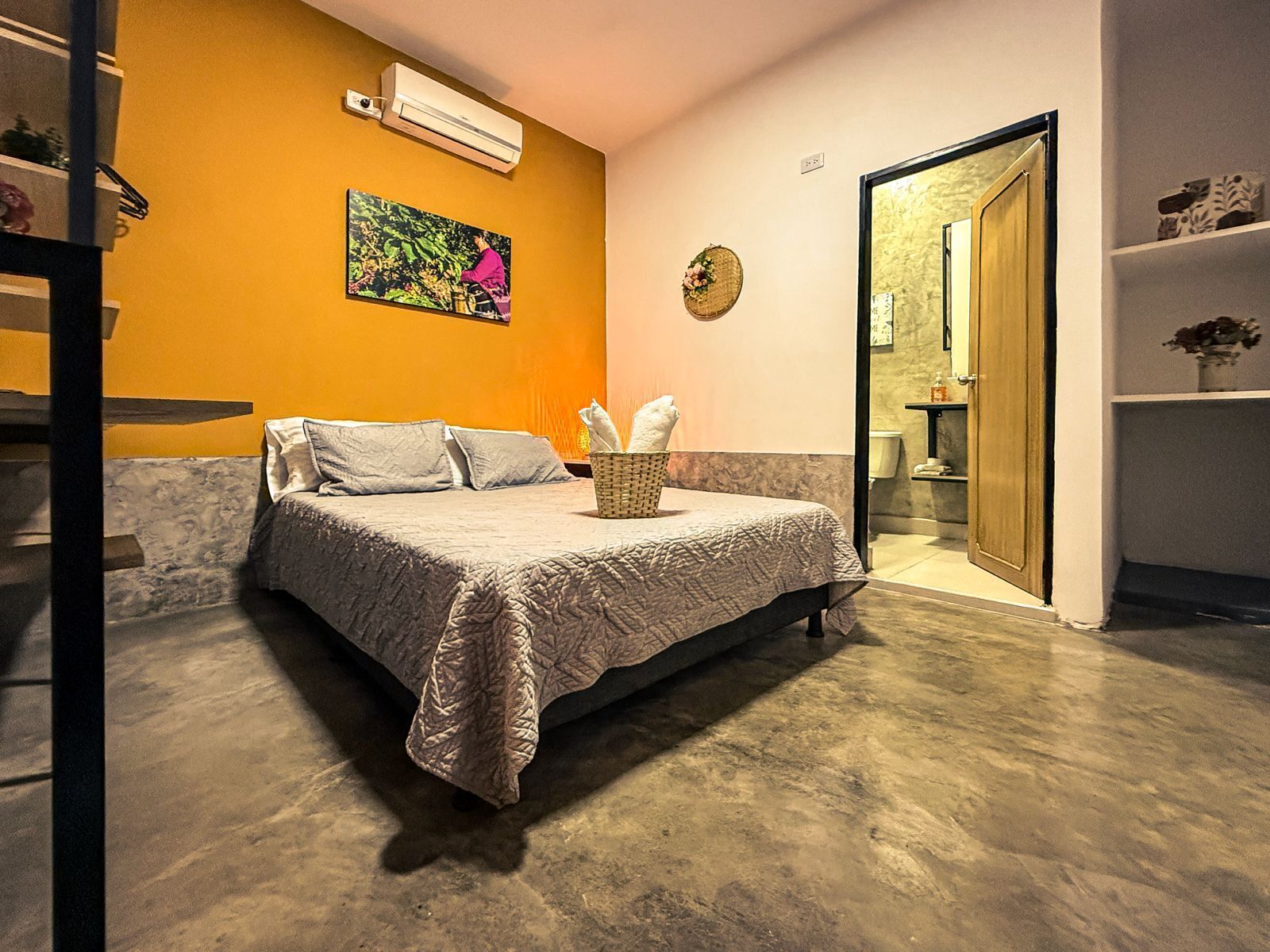 A bedroom with a bed, a wall-mounted air conditioner, a painting, a clock, and a door leading to a bathroom.