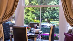 A dining area with a table set for two, a window with curtains, and a view of trees and a car outside. - Maida Vale - Carlton Vale Apartments (London)