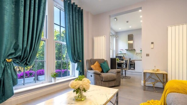 A modern living room with a marble coffee table, a sofa, and a chair with cushions. There is a view of a kitchen and dining area in the background. - Maida Vale - Carlton Vale Apartments (London)