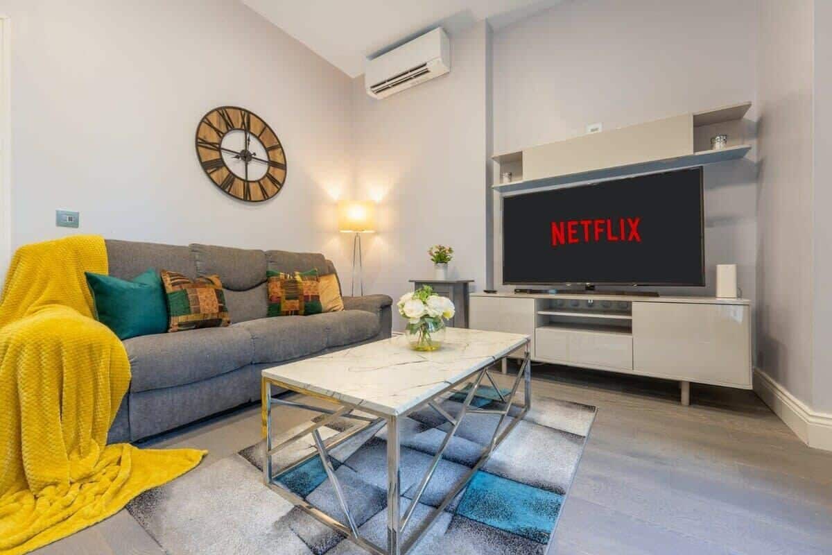 A living room with a grey sofa, a yellow blanket, a clock, a TV displaying 'NETFLIX', and a modern coffee table.