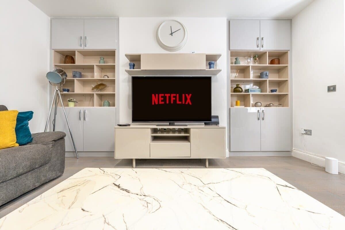 A modern living room with a flat-screen TV mounted on a white cabinet, a clock above it, and a bookshelf with various items.