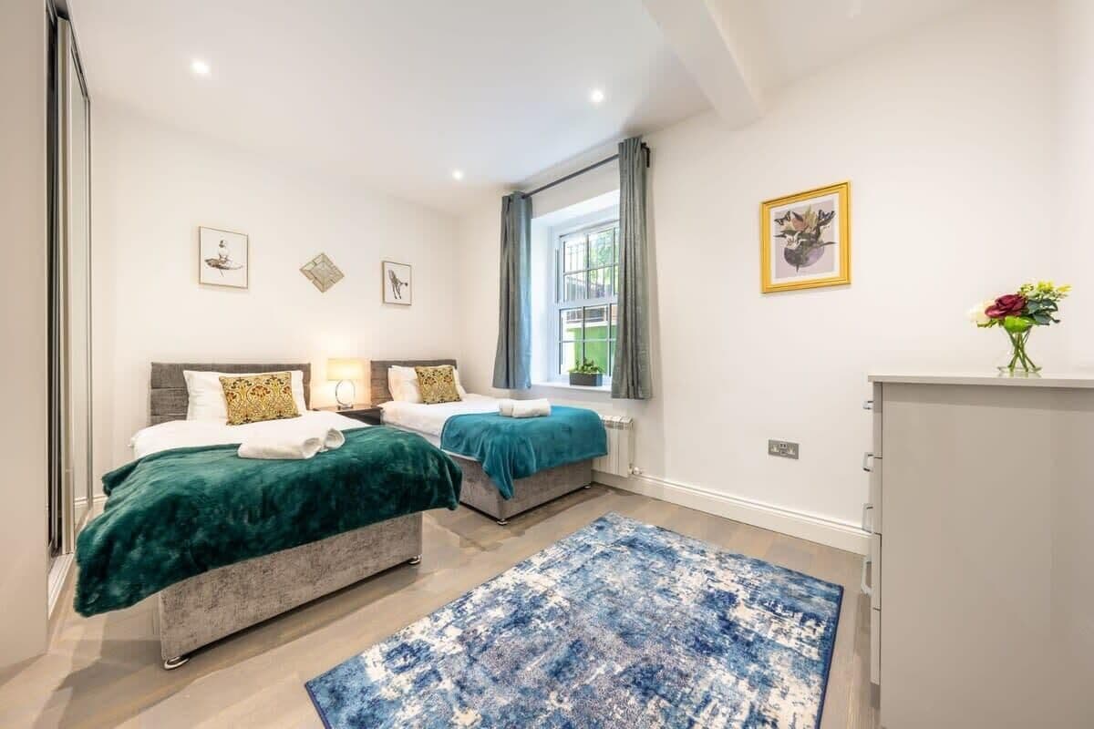 Een slaapkamer met twee bedden, een raam met gordijnen, een bureau met een vaas en een blauw vloerkleed.